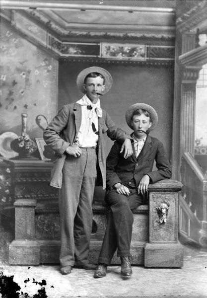 Full-length studio portrait in front of a painted backdrop of two young men wearing hats and suits. The man on the left is posing standing with a cigar in his mouth, and the man on the right is posing sitting with a pipe in his mouth.