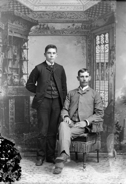 Full-length studio portrait in front of a painted backdrop of a man posing standing on the left, and a man with a moustache posing sitting in a fringed chair on the right.