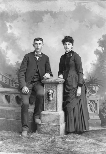 Full-length studio portrait in front of a painted backdrop of a man and woman posing sitting and standing on the right next to a prop stone wall and column.