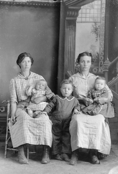 Full-length studio portrait in front of a painted backdrop of two woman posing sitting. They are each holding an infant wearing clothing with the same pattern as their dresses. In the center is a boy posing standing with his hands on the knees of each woman.