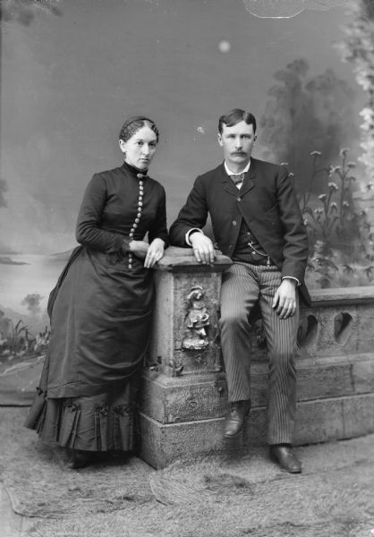 Full-length studio portrait in front of a painted backdrop of a woman standing and leaning on a prop stone column, and a man sitting on a prop stone wall. 