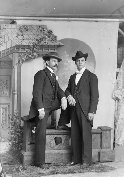 Full-length studio portrait of two men wearing suits and hats, posing standing in front of a painted backdrop. The man on the left has his left leg up on a low prop stone wall.
