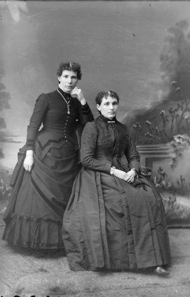 Full-length studio portrait of two woman posing in front of a painted backdrop. The woman on the left is standing with her elbow propped on the back of the chair the woman in front of her is sitting on.