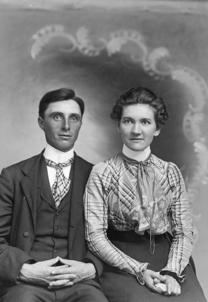 Three-quarter length studio portrait of a man and woman posing sitting in front of a painted backdrop.