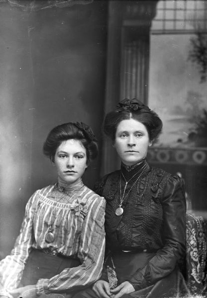 Three-quarter length studio portrait of two woman posing sitting together in front of a painted backdrop. Both women are wearing watches or lockets on chains.