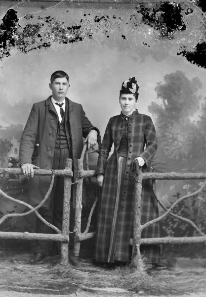 Full-length studio portrait in front of a painted backdrop of a man and woman posing standing with a prop fence and gate. The woman is wearing a long, plaid coat and a hat. The man is wearing a three-quarter length coat over his suit.