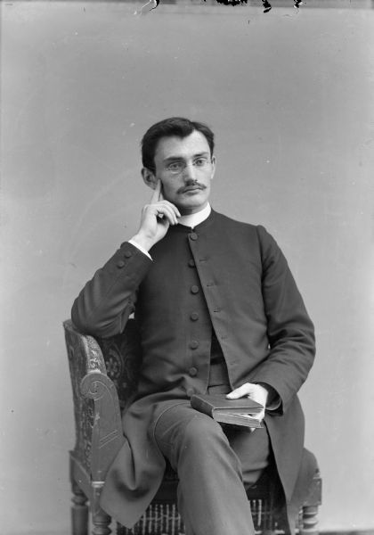 Three-quarter length studio portrait of a man with a moustache, and wearing eyeglasses, posing sitting and holding a book on his lap.