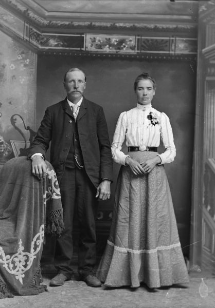 Full-length studio portrait in front of a painted backdrop of a man and woman posing standing together. The man has his hand on a cloth draped chair. The woman is wearing a corsage.