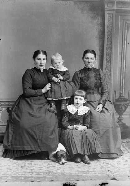 Full-length studio group portrait in front of a painted backdrop of two women posing sitting and flanking a girl, a child, and a dog lying on the carpet.