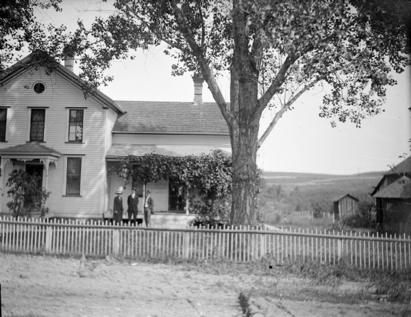 View from street of a wooden house with a front porch covered with a vine. Three people are posing standing in the yard behind a wooden fence. There are buildings on the far right, and a valley and hills in the background.