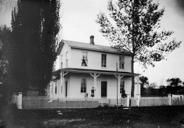 View across yard towards the southwest corner of South 2nd and Johnson (Grove). Owned by Johnson. There is a man standing inside the fence, and a girl standing on the porch. A woman holding a baby is looking out of a second-story window.