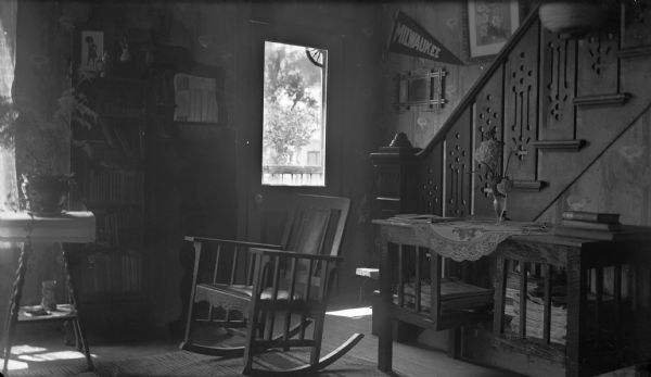 Interior view of a home towards the front door with large window, and the base of a staircase on the right. In the center is a rocking chair, and a table and plant on the left near a window. Identified as the home of Jane Thomas in Spring Creek.