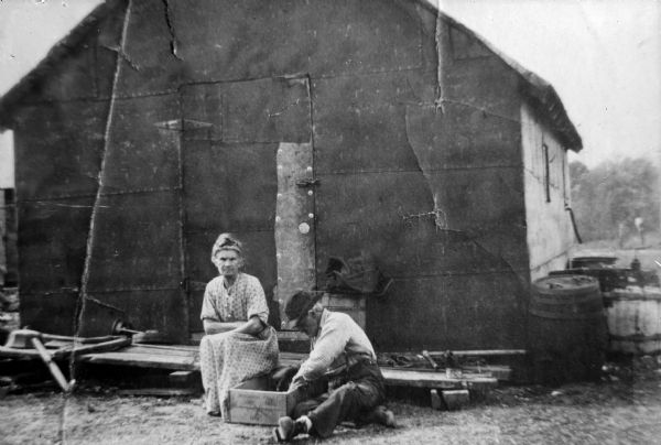 Copy photograph of an exterior view of a wooden house with a European American man and European American woman posing sitting in the yard.