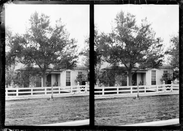 Stereograph of an exterior view of a wooden dwelling. A couple is sitting on the porch, and a man is standing just inside the fence. A dog is lying next to hitching post in the street.