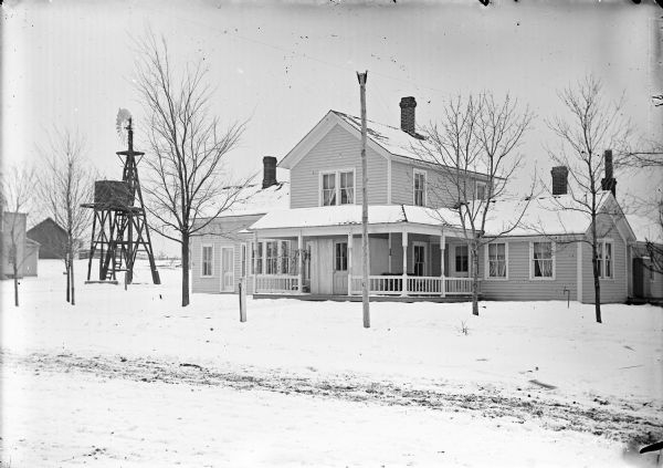 Exterior view from street of a two-story wooden house, with a windmill on the left. There is snow on the ground.