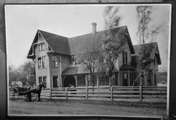 Copy photograph of an exterior view of a two-story wooden dwelling. A horse and buggy are parked in the street in front of a fence. A woman and two children are sitting on the porch.