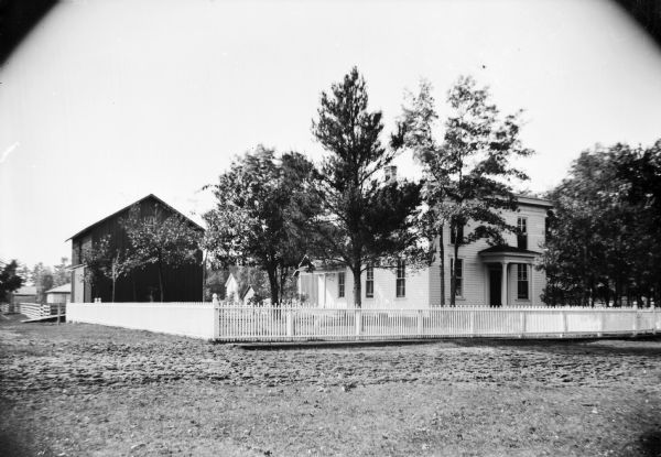 Exterior view from street of a two-story wooden house with a surrounding picket fence. There are farm buildings in the background.