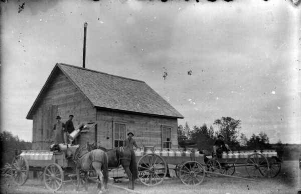 Exterior view of a wooden creamery building, with men and horse-drawn wagons transporting milk cans.