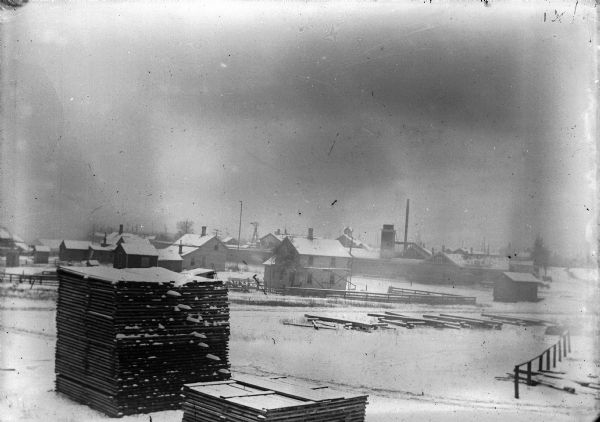 Exterior view of snow-covered wooden buildings. Stacks of wood and a railroad track crossing is in the foreground.