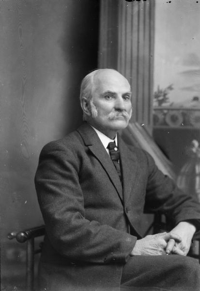 Studio portrait in front of a painted backdrop of an elderly European American man posing sitting. Identified as Charles J. Van Schaick.