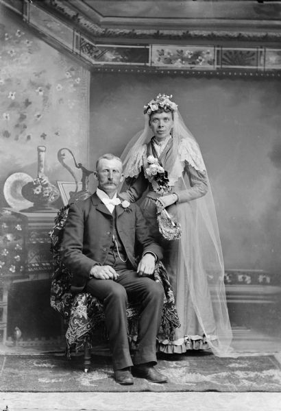Full-length studio portrait in front of a painted backdrop of a middle-aged wedding couple, bride and groom. The man is posing sitting on the left, and a the woman is posing standing on the right.