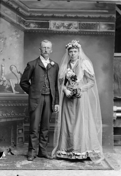 Full-length studio portrait of a middle-aged wedding couple posing in front of a painted backdrop. The man is posing standing on the left, and the woman is posing standing on the right.