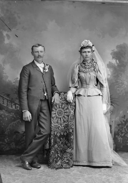 Full-length studio portrait of a wedding couple, bride and groom, posing in front of a painted backdrop. The man is standing on the left, and the woman is standing on the right.