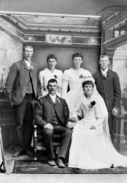 Studio group portrait of a wedding group, all wearing corsages, posing in front of a painted backdrop. includes the bride and groom, a man sitting on the left, and a European American woman sitting on the right. Behind them two men are standing and flanking two European American women who are also standing.