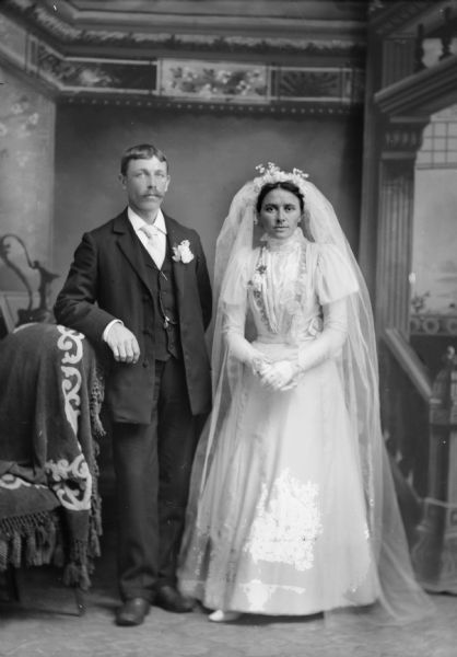 Studio portrait of a wedding couple, bride and groom, posing in front of a painted backdrop. The man is standing on the left, and the woman is standing on the right.
