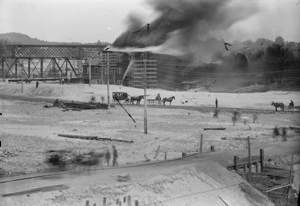 Elevated view of the fire at the McGillivray factory. Horse-drawn vehicles and onlookers are on the road in the foreground.