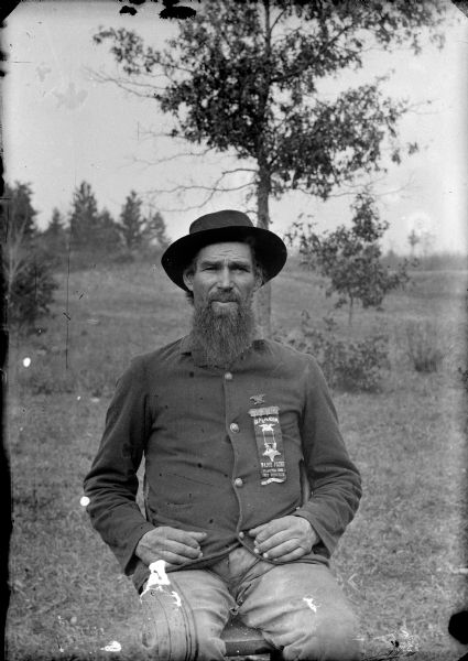 Three-quarter length outdoor portrait of a man with a moustache and long beard posing sitting, probably a Civil War veteran and member of the Grand Army of the Republic. The ribbon on his jacket reads "In Memorium, Major Paine, Post No. 166, Dep T Wisconsin."