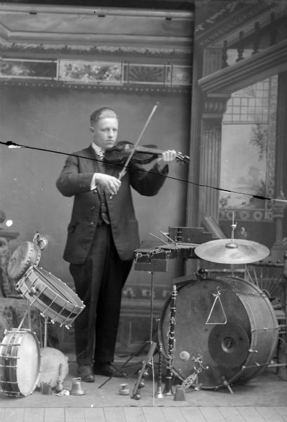 Full-length studio portrait in front of a painted backdrop of a European American man posing standing and playing a violin among other musical instruments set up around him.