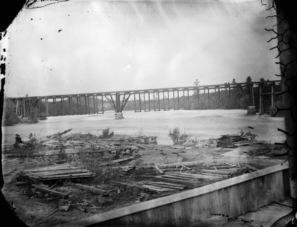 View across water towards what is identified as the second railroad bridge at Black River Falls. Note the wood trestle on the right.