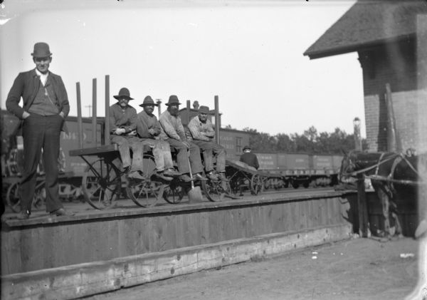 Exterior view of work men posing sitting on a wagon on the platform at the railroad station.