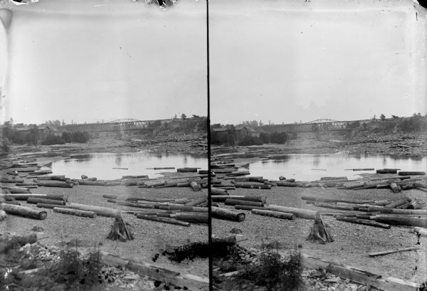 Outdoor view, in stereograph form, looking up the Black River from Black River Falls. There are logs in the river in the foreground, and a bridge in the background.