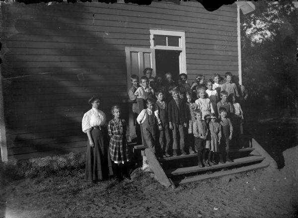 Group portrait of a teacher and school children posing standing outside a wooden school house.