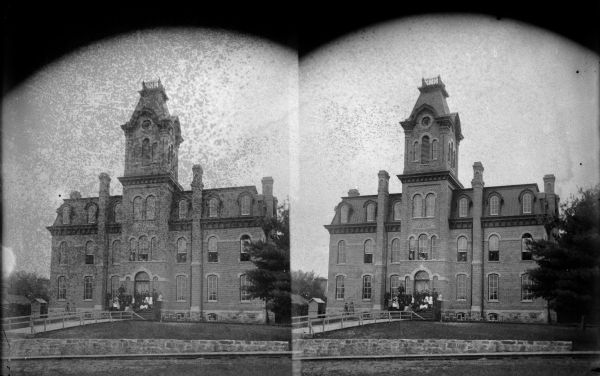 Outdoor view, in stereograph form, of the elementary school in Black River Falls. A group of faculty are gathered on the stairs and in the second story windows.