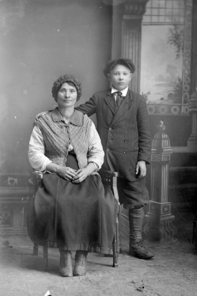 Studio portrait in front of a painted backdrop of a woman posing sitting on the left and a boy standing next to her on the right. The boy is wearing a suit, and the woman is wearing a knit vest.