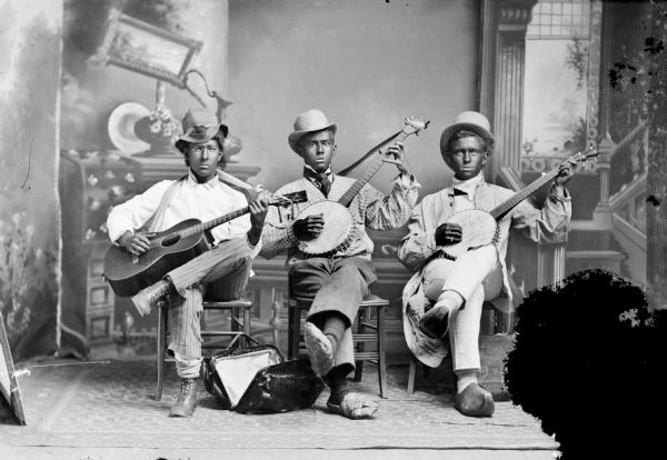 Studio portrait in front of a painted backdrop of three European American men, posing sitting. The men are wearing blackface make-up, and playing a guitar and banjos.