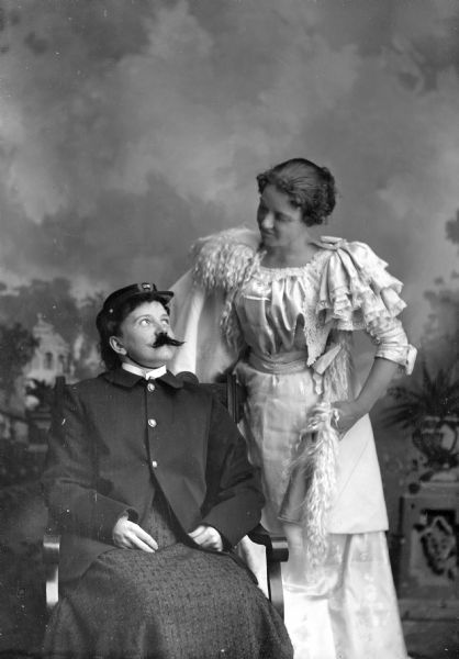 Studio portrait in front of a painted backdrop of a woman posing sitting on the left wearing a false moustache, uniform jacket and hat, and a woman wearing a white dress standing next to her on the right.