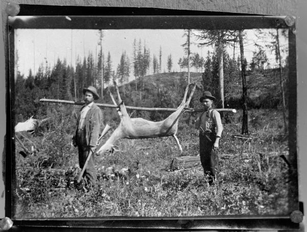 Outdoor portrait of two men with a deer suspended from a wood pole the men are carrying on their shoulders.