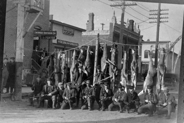 Copy photograph of an outdoor portrait of a group of men standing and kneeling in front of a line of deer suspended across First Street in Black River Falls, probably between 1908-1911.