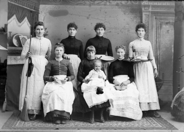 Studio portrait in front of a painted backdrop of seven European American women. Four of them are posing standing and three are posing sitting, with an infant sitting in the lap of the women in the center. All the women are holding household implements. Identified as probably Swedish maids.