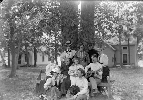 Outdoor group portrait of ten people posing sitting and standing in front of a large tree, with houses in the background. One man is holding a guitar, and a man in the center has a dog sitting in his lap.
