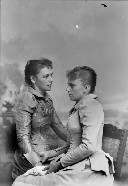 Waist-up studio portrait in front of a painted backdrop of two unidentified women posing sitting and facing each other.