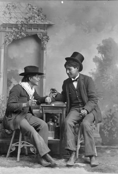 Studio portrait in front of a painted backdrop of two Native American men posing sitting and holding the same glass over a table.