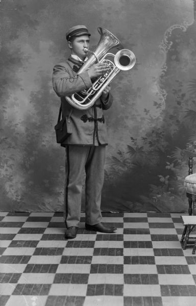 Studio portrait in front of a painted backdrop of a European American man posing standing and playing a Double Bell Euphonium.