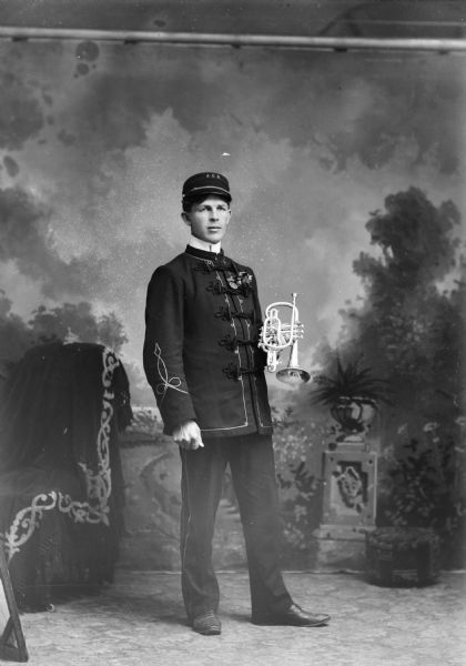 Studio portrait of a European American man posing standing and holding a trumpet. Identified as possibly a member of the Alma Center Band.