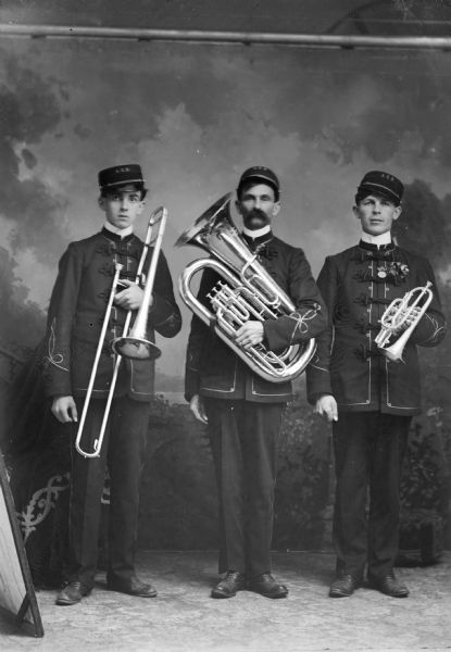 Studio group portrait in front of a painted backdrop of three European American men posing standing. Each man is holding, from left to right, a trombone, euphonium and cornet. Identified as possibly members of the Alma Center Band.