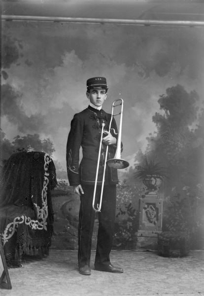 Studio portrait of a European American man posing standing and holding a trombone. Identified as possibly a member of the Alma Center Band.